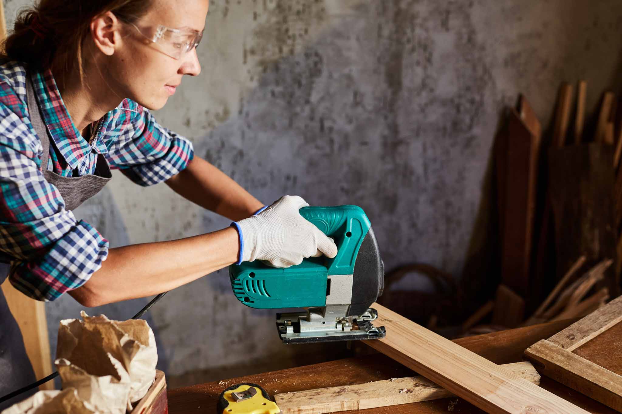 Female Carpenter sawing wood with an electric jigsaw. Woman in apron doing some carpentry work in his workshop, using electric jigsaw.